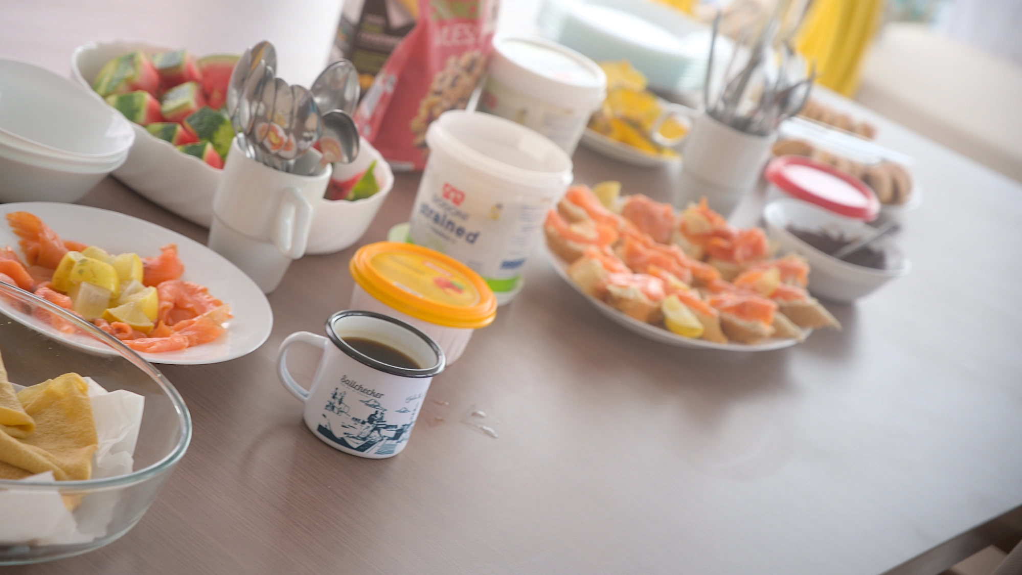 Breakfast table with smoked salmon, fresh watermelon, bread with toppings, yoghurt, chocolate spread, and a SailChecker coffee mug